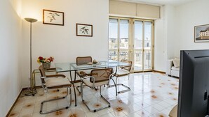 Un salon moderne avec une table à manger en verre, des chaises en métal, un canapé, un téléviseur et un balcon offrant une vue sur la ville.