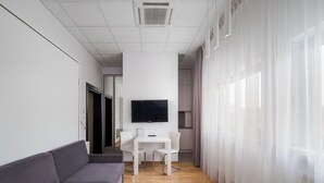 A modern hotel room with a grey sofa, a small white table, a flat-screen TV mounted on the wall, and sheer curtains on the windows.