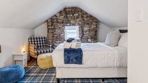 A cozy bedroom with a stone wall, a checkered chair, a bed with a blue blanket, and a small bedside table with a lamp. - Charming Blake Cottage | Augusta Wine Country (Augusta)