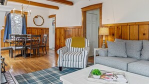 A cozy living room with wooden beams, a striped armchair, a sofa, and a dining area with a table and chairs. - Charming Blake Cottage | Augusta Wine Country (Augusta)