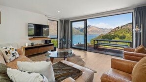 A modern living room with a large TV, leather sofas, a glass coffee table, and a scenic mountain view through large windows. - Sam`s Sanctuary | Stunning Views & Spacious Home (Queenstown)