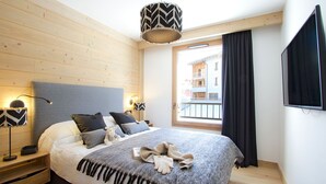 Un dormitorio con cama, lámparas de noche, ventana con vista a edificios y televisor montado en la pared.