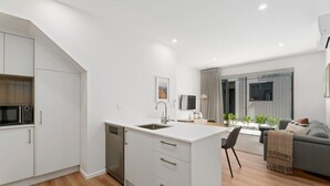A modern kitchen with white cabinetry, a built-in dishwasher, and a small island with a sink. There is a dining area with a gray sofa and a coffee table. - Skippers Rest | Close to ski fields & parking (Queenstown)