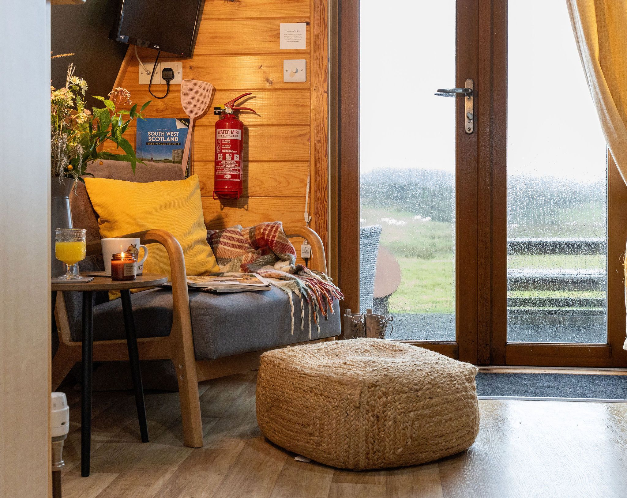 Ein gemütliches Zimmer mit Holzvertäfelung, einem Bett mit karierter Decke, einem kleinen Tisch mit Tasse und Glas sowie einem Feuerlöscher an der Wand.