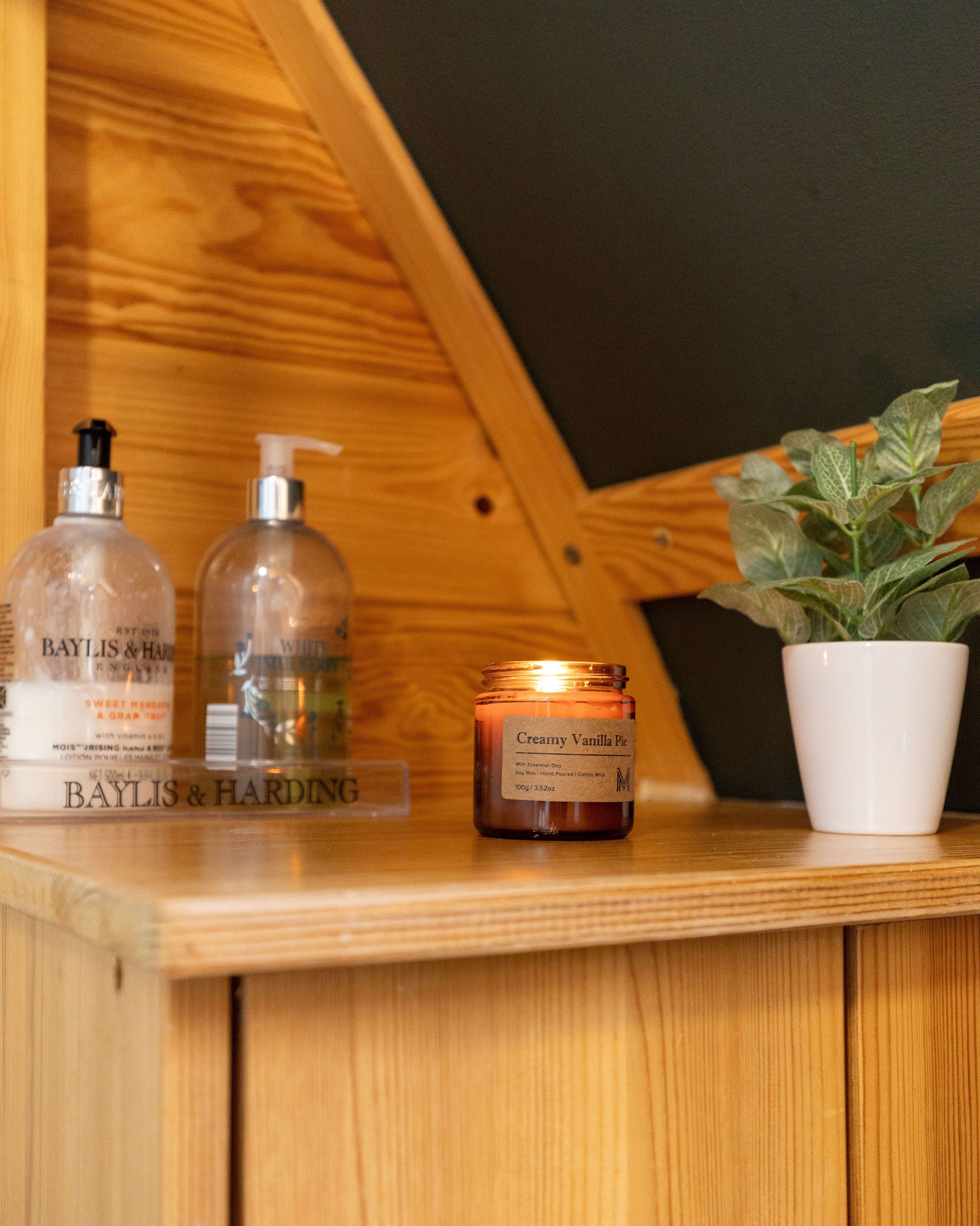 Une étagère en bois sur laquelle sont posées trois bouteilles de produits Baylis & Harding, un pot de crème dessert à la vanille et une plante en pot.