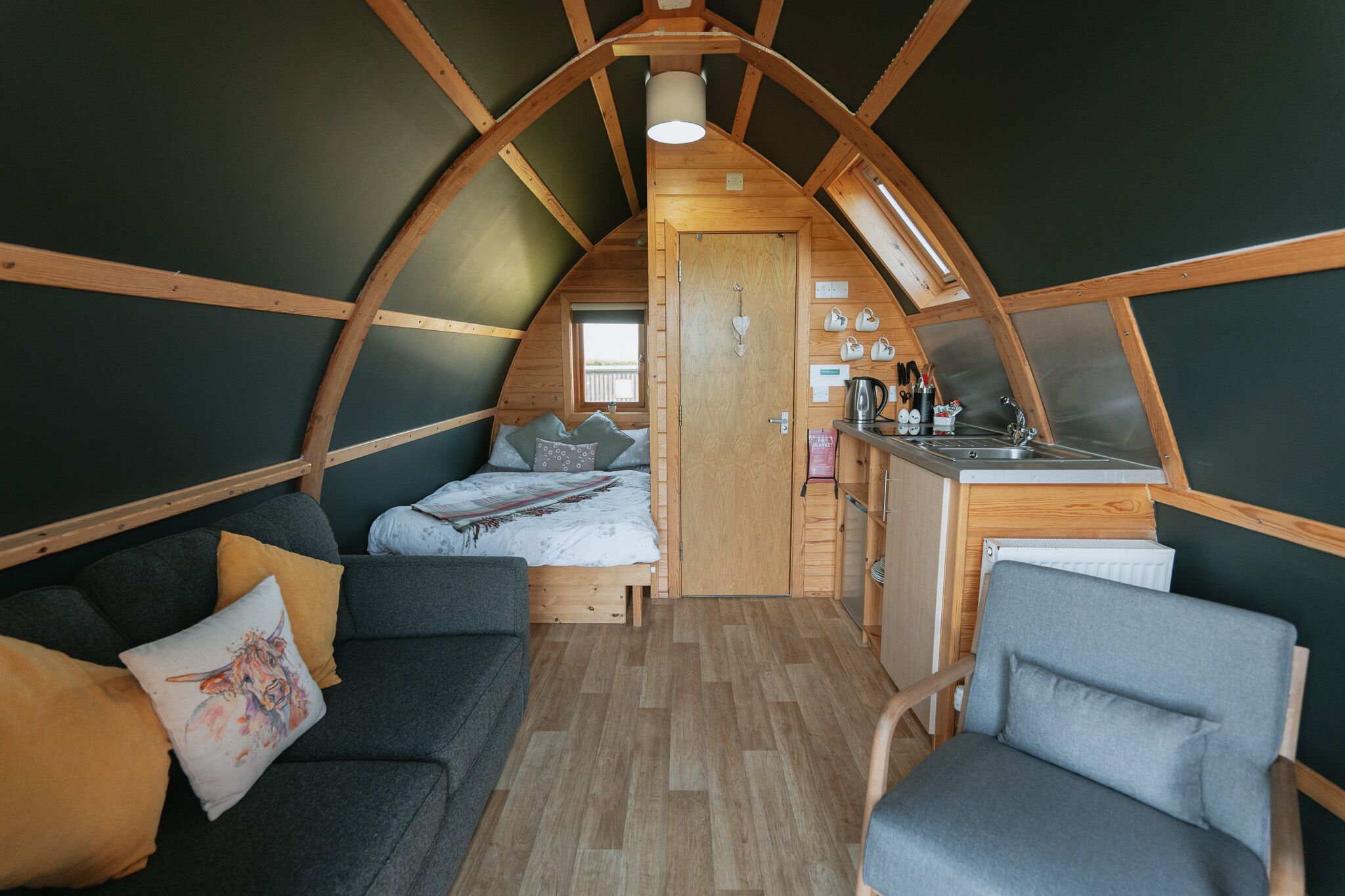 Une chambre de style loft, à la fois chaleureuse et partiellement ouverte, comprenant un lit, un canapé et un fauteuil. On peut apercevoir une petite cuisine équipée d’un évier et d’une cuisinière.
