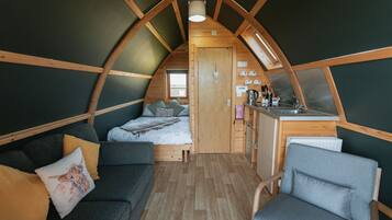 Une chambre de style loft, à la fois chaleureuse et partiellement ouverte, comprenant un lit, un canapé et un fauteuil. On peut apercevoir une petite cuisine équipée d’un évier et d’une cuisinière.