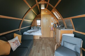 Cómodo dormitorio tipo loft, parcialmente abierto, con cama, sofá y silla. Se observa una pequeña cocina con lavaplatos y estufa.