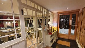 Un couloir avec un espace cuisine visible à travers une cloison en verre, une porte en bois ornée d’un motif représentant un paysage enneigé et un sol carrelé.