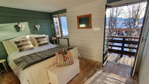 Une chambre avec un lit, une petite commode blanche, un sac coloré, une fenêtre et une vue sur les montagnes.