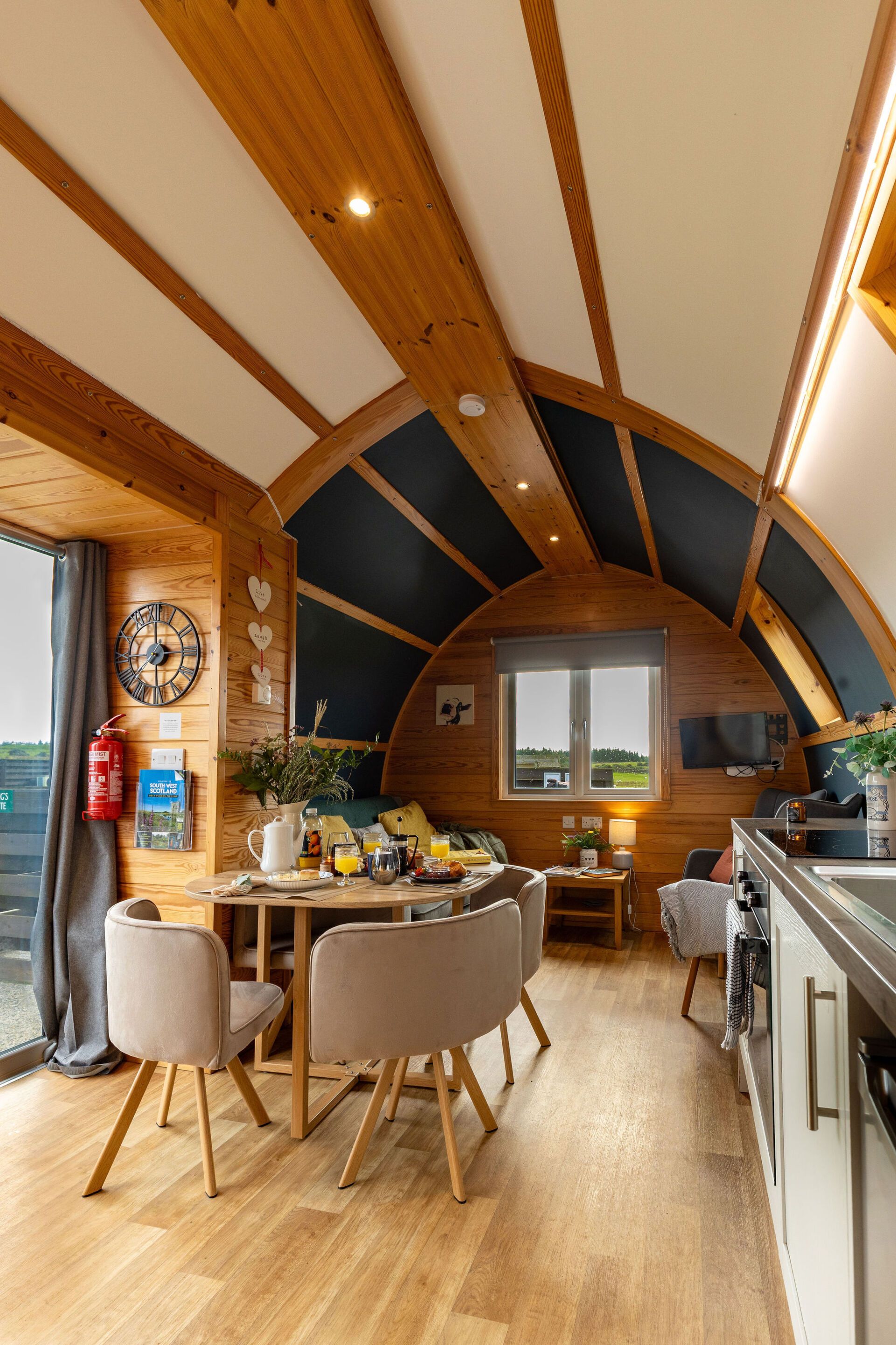 A cozy interior with a wooden ceiling, a dining area with chairs, a table with breakfast items, and a kitchen area.