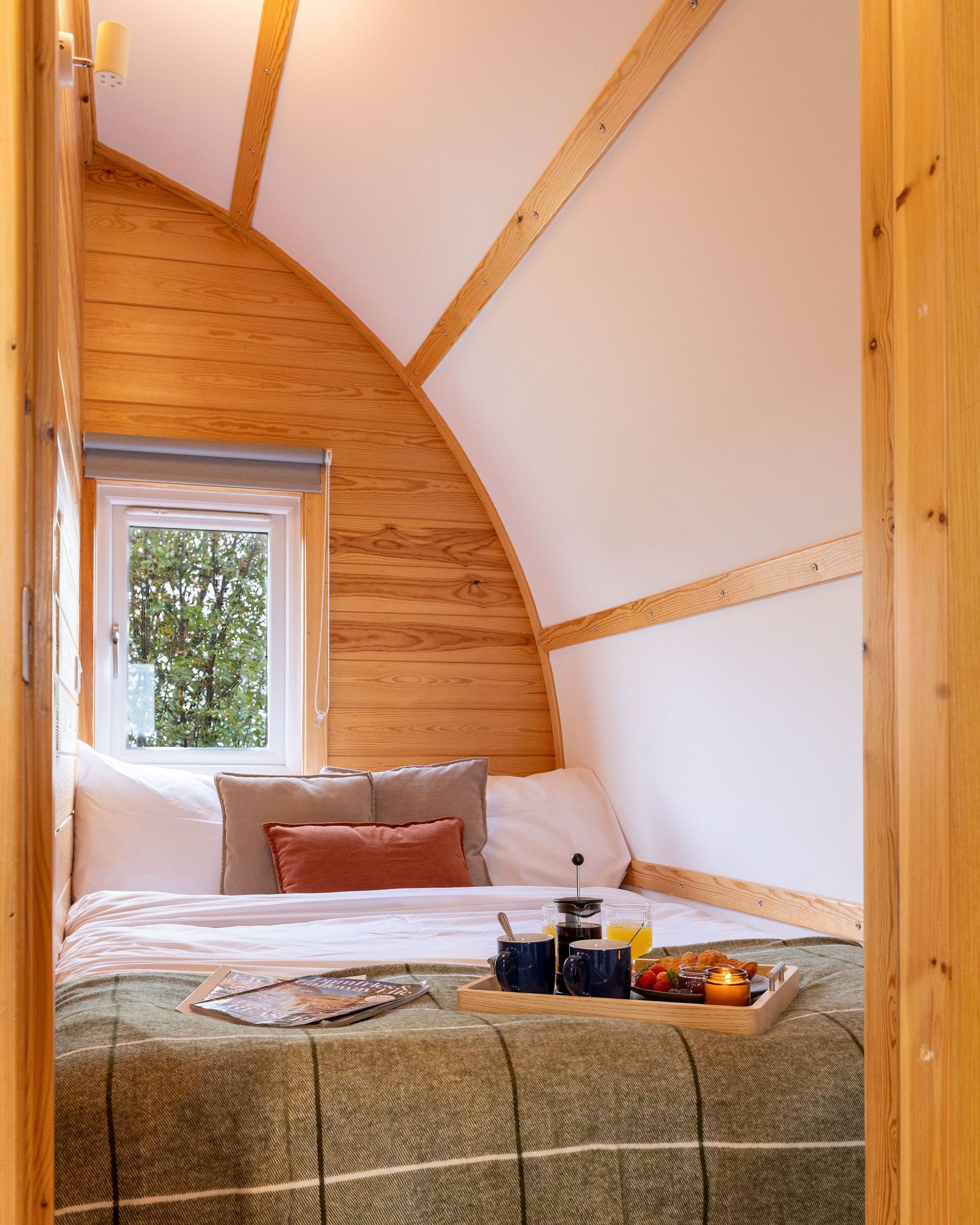 Ein gemütliches Schlafzimmer mit gewölbter Decke, einem Fenster, einem Bett mit Kissen, einem Nachttisch mit Frühstücksgebäck und Blick ins Grüne.