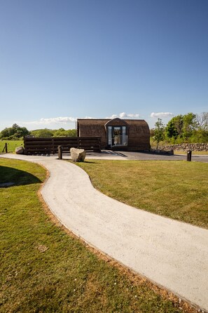 A rustic wooden building with a large door and a small balcony, surrounded by a paved path and grassy areas. - Luxury Rural Cabin With Ensuite & Hot Tub (Newton Stewart)