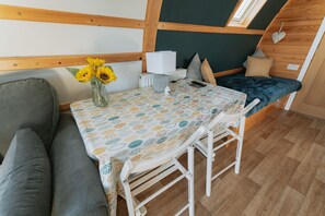 Ein gemütliches Zimmer mit Holzbalkendecke, einem Bett mit dunkelblauer Bettdecke, einem kleinen Tisch mit gemustertem Tischläufer, einer Vase mit Sonnenblumen und einem grauen Sofa.