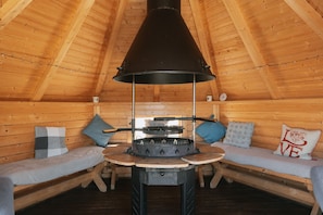 A cozy wooden cabin with a central wood stove, two wooden benches with cushions, and a pitched wooden ceiling.