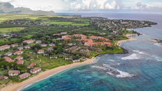 An aerial view of a coastal resort with beachfront properties, a golf course, and a mountainous backdrop.