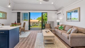 A spacious living area with a sofa, coffee table, and a dining area with chairs. There is a sliding glass door leading to an outdoor balcony with a view of palm trees.