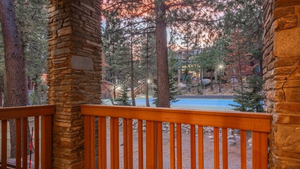 A wooden deck with stone pillars, overlooking a pool and a forested area with a building in the background.