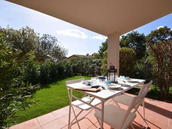 An outdoor patio with a white table and chairs, surrounded by greenery and a clear sky.