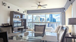 Un salón moderno con una mesa de centro de vidrio, asientos cómodos, un televisor y vistas a la ciudad a través de grandes ventanales.