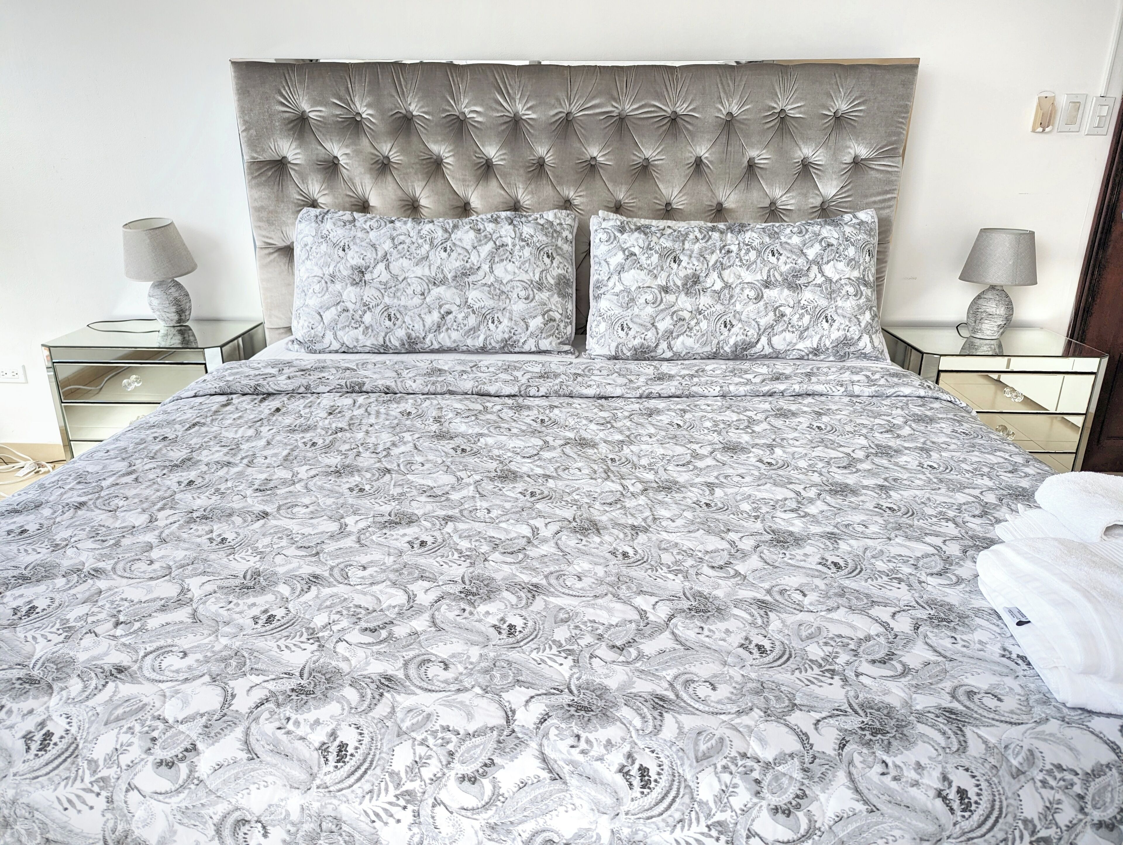 Een slaapkamer met een gestuikt hoofdeinde, twee bijpassende kussens met een patroon, een bed met een bedsprei met paisleyprint en twee nachtkastjes met lampen.