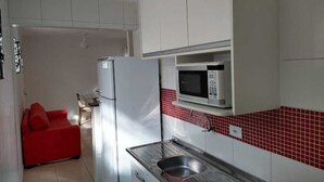 Une cuisine de taille réduite avec des meubles blancs, un plan de travail recouvert de carreaux rouges, un four à micro-ondes et un évier. Un canapé rouge se trouve dans le salon attenant.