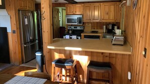A compact kitchen with wooden cabinets, a stainless steel refrigerator, a microwave, a stove, and a coffee maker. There are two stools at a wooden bar counter.