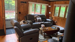 A cozy cabin interior with a wooden interior, a leather sofa, a dining table, and a large window overlooking a deck.