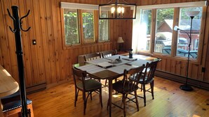 A wooden-paneled dining room with a table set for four, a chandelier, and large windows offering a view of the outdoors.