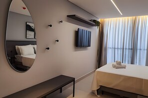 A modern hotel room with a large mirror, a bedside table, a bed with a white cover, a small shelf, and a view of the outdoors through sheer curtains. - Alta Garden (João Pessoa)