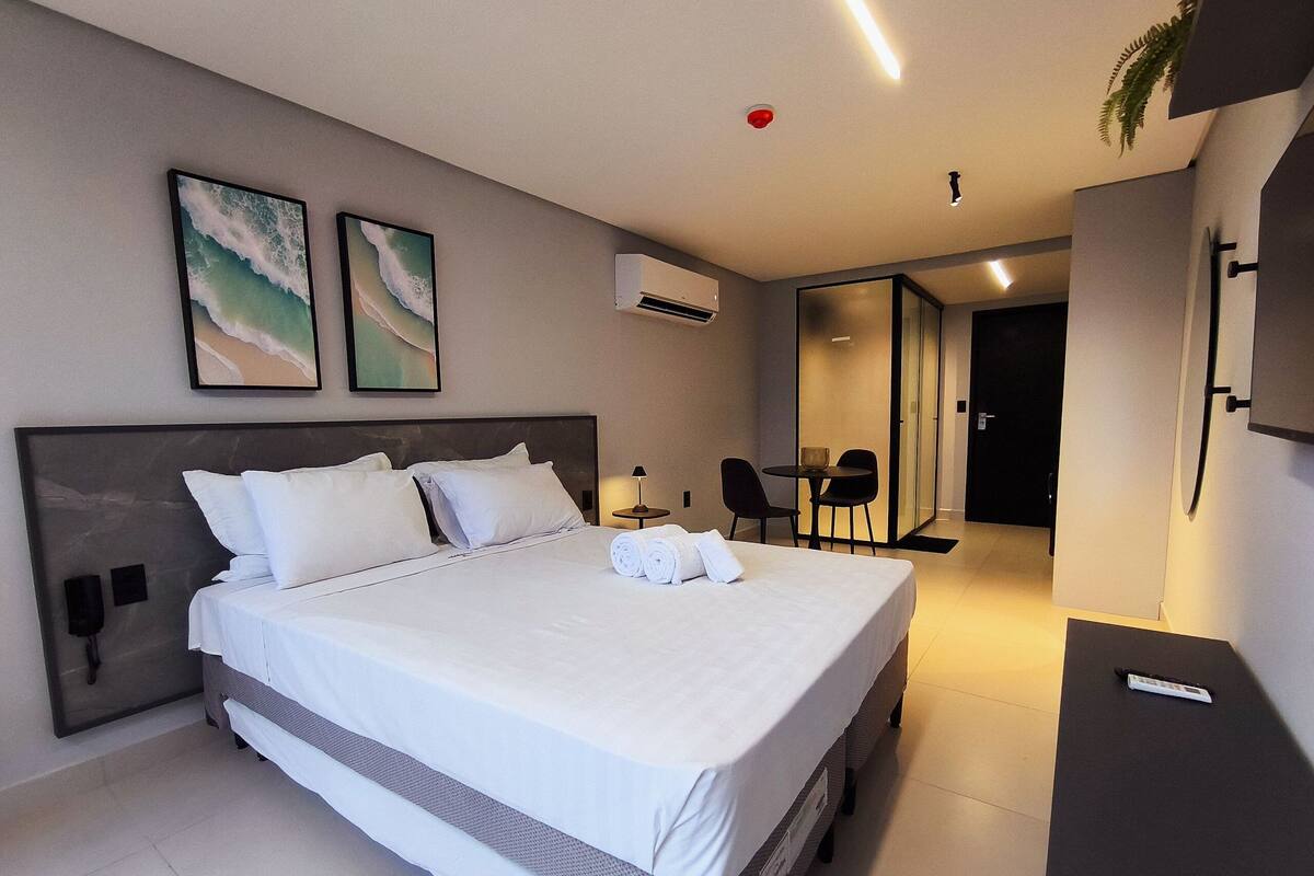 Quarto de hotel moderno com uma cama grande, duas unidades de ar condicionado fixadas na parede, uma pequena mesa com duas cadeiras e uma porta que dá para um banheiro.