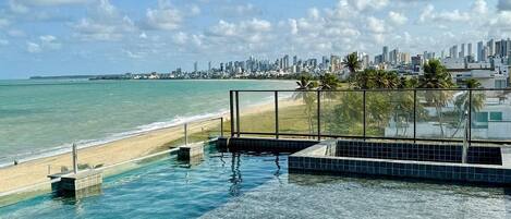 A rooftop infinity pool with clear water, a city skyline in the background, and a beach.