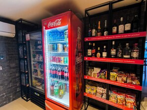 A vending machine with Coca-Cola and various snacks, and a shelf with alcohol bottles. - Alta Garden (João Pessoa)