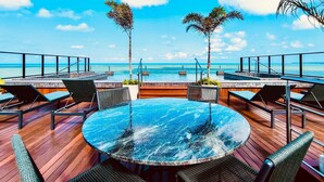 Mesa redonda con cubierta de vidrio rodeada de sillas, con vistas a una piscina y una playa.