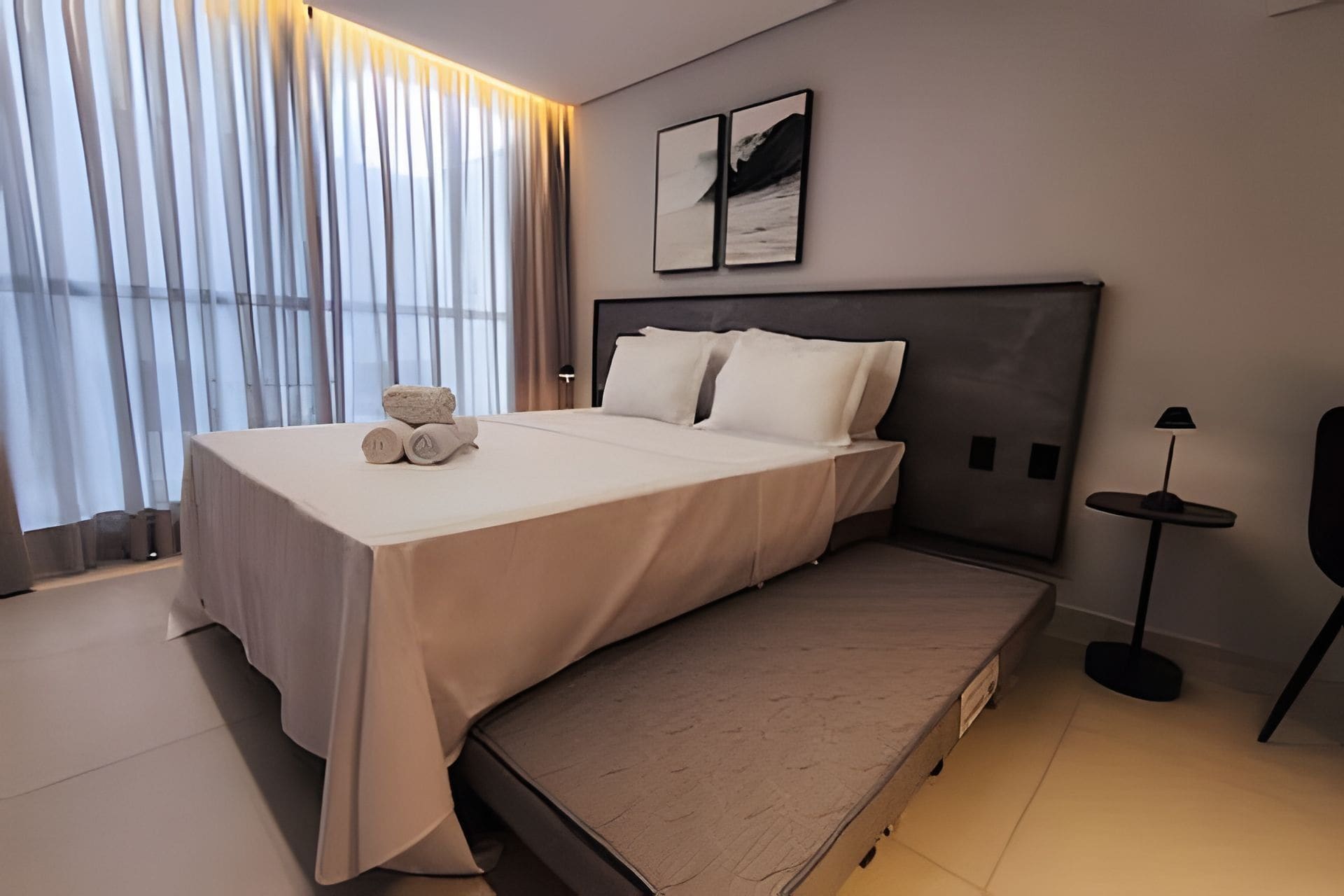 Ein modernes Hotelzimmer mit einem großen Bett, einem Nachttisch, einer Lampe und Blick auf die Stadt durch große Fenster.