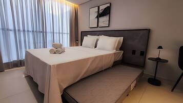 Ein modernes Hotelzimmer mit einem großen Bett, einem Nachttisch, einer Lampe und Blick auf die Stadt durch große Fenster.