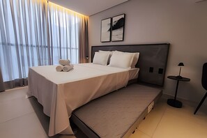 A modern hotel room with a large bed, a bedside table, a lamp, and a view of the city through large windows. - Alta Garden (João Pessoa)