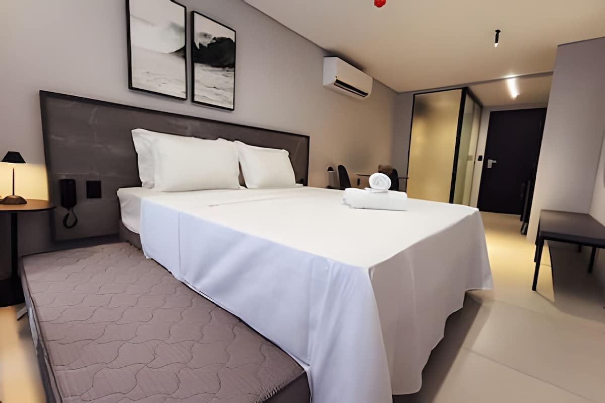 Quarto de hotel moderno com uma cama grande, mesa de cabeceira com abajur, telefone e ar condicionado fixado na parede.