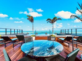 Mesa redonda com tampo de vidro, rodeada por cadeiras, com vista para uma piscina e uma praia.