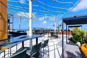 An outdoor seating area with tables and chairs, a covered section with a view of the ocean, and a wooden structure on the left. - Alta Garden (João Pessoa)