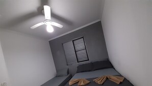 Een knus kamertje met een bed, een bankje, een plafondventilator en een raam met jaloezieën.