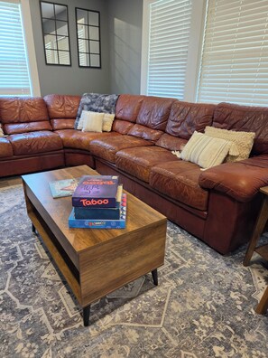 A living room with a brown leather sectional sofa, a wooden coffee table with board games, and patterned carpet.