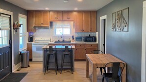 A modern kitchen with wooden cabinets, a central island with bar stools, a dining table with chairs, and a microwave. - Cozy 3-bedroom house minutes to Okemo and downtown Ludlow (Mount Holly)
