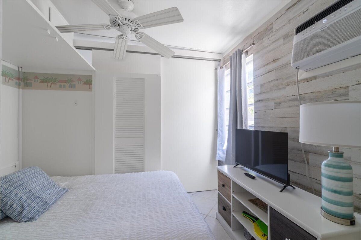 Een slaapkamer met een bed, een plafondventilator, een televisie op een standaard en een raam met gordijnen.