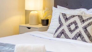 Una cama bien hecha con un cojín estampado, una lámpara de noche y un pequeño objeto decorativo en la mesita.