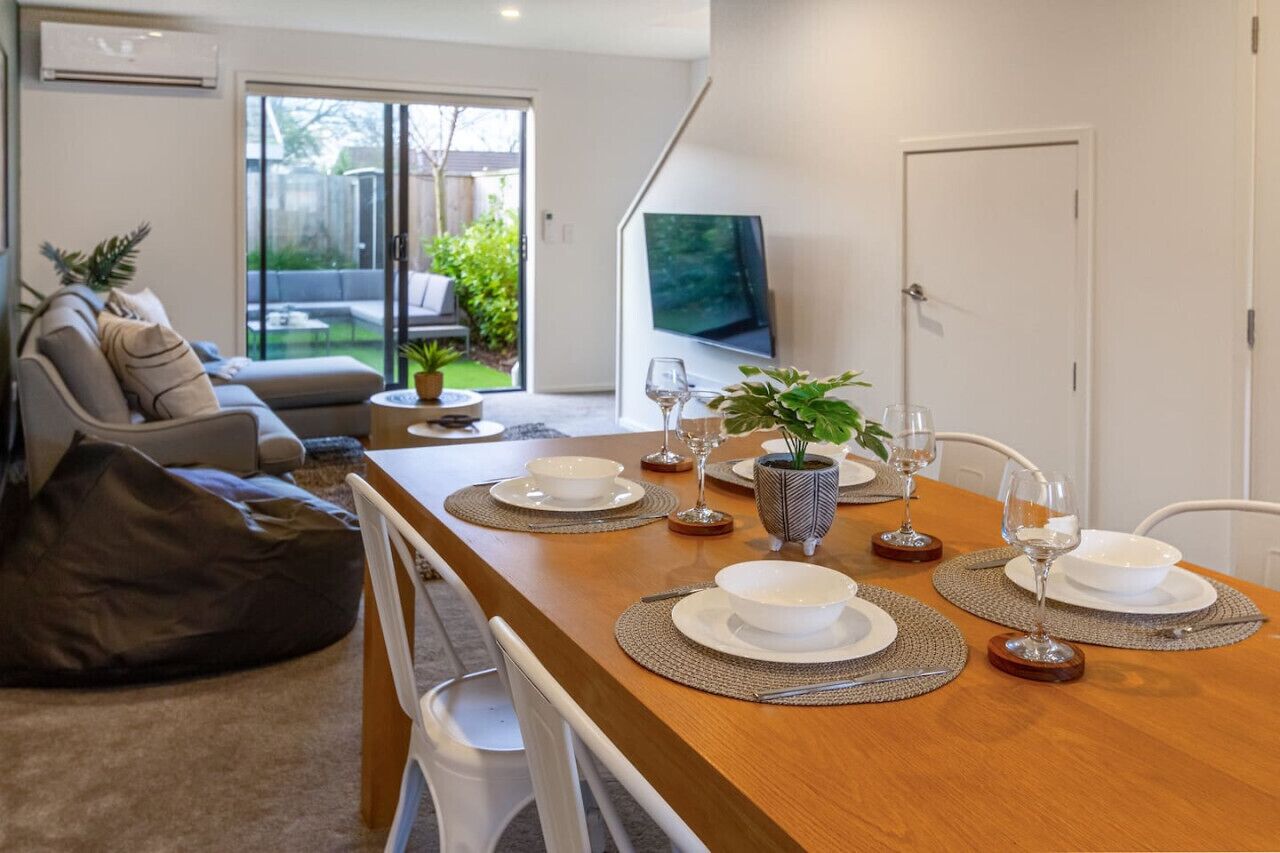 A modern living room with a dining table set for four, a sofa, a TV, and a sliding glass door to a backyard.