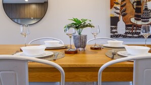 Una mesa de comedor dispuesta con cuencos, platos y cubiertos blancos, flanqueada por sillas blancas. Una planta en maceta y un jarrón decorativo se encuentran sobre la mesa.
