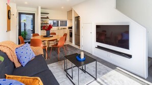 A modern living room with a sofa, coffee table, and TV. There is a dining area with chairs and a table. The kitchen is visible in the background. - Bright and Stylish 2BR Near Addington Park (Christchurch)
