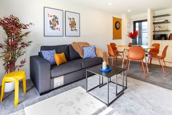A modern living room with a grey sofa, orange chairs, and a coffee table. There are framed artwork on the wall and a plant in the corner.