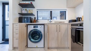 A compact kitchen with a built-in washing machine, wooden cabinets, and a window providing natural light. - Bright and Stylish 2BR Near Addington Park (Christchurch)